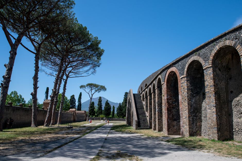 Explore the ancient ruins of Pompeii under clear blue skies with Mount Vesuvius visible in the distance.